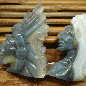 Puede incluir: Dos esculturas talladas en piedra de &aacute;gata de jefes nativos americanos, una con un tocado de plumas y la otra con una trenza larga. Las esculturas son de color gris claro con acentos blancos y morados.