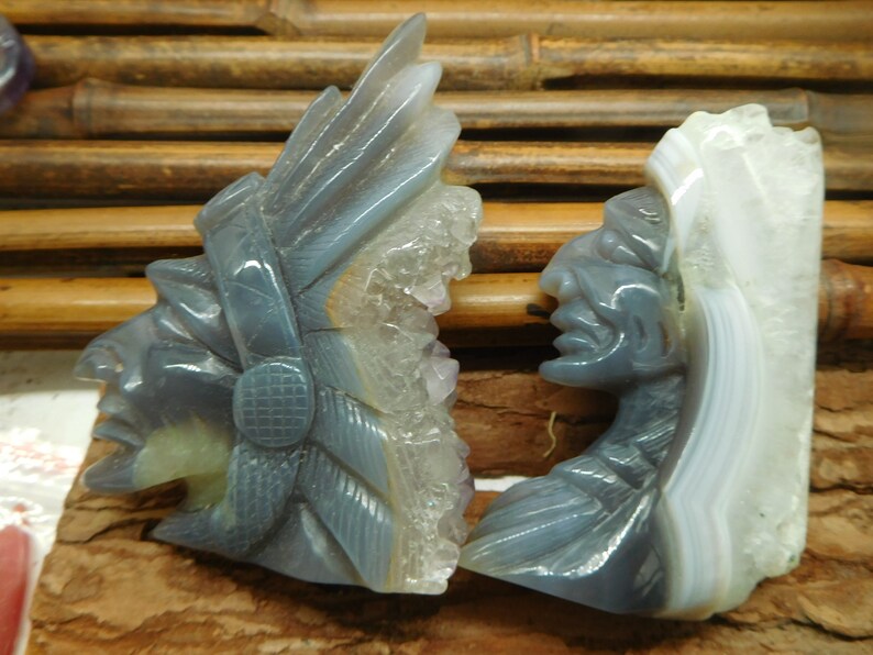 Puede incluir: Dos esculturas talladas en piedra de &aacute;gata de jefes nativos americanos, una con un tocado de plumas y la otra con una trenza larga. Las esculturas son de color gris claro con acentos blancos y morados.