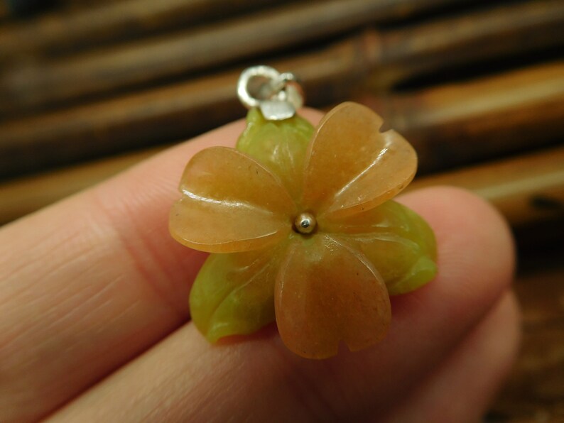 Puede incluir: Un delicado colgante de flor hecho de piedra verde y naranja tallada. El colgante tiene un aro de plata y una peque&ntilde;a perla de plata en el centro.