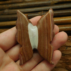 May include: A brown and white butterfly-shaped stone with red and white stripes. The stone is carved in the shape of a butterfly with its wings spread out.