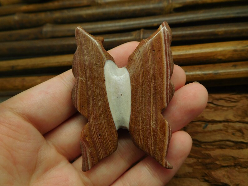 May include: A brown and white butterfly-shaped stone with red and white stripes. The stone is carved in the shape of a butterfly with its wings spread out.