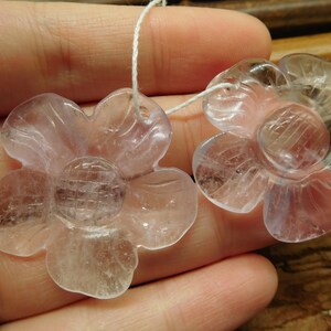 May include: Two clear, translucent flower-shaped pendants made of amethyst. The flowers have five petals and a center with a grid pattern. The pendants are strung on a white cord.