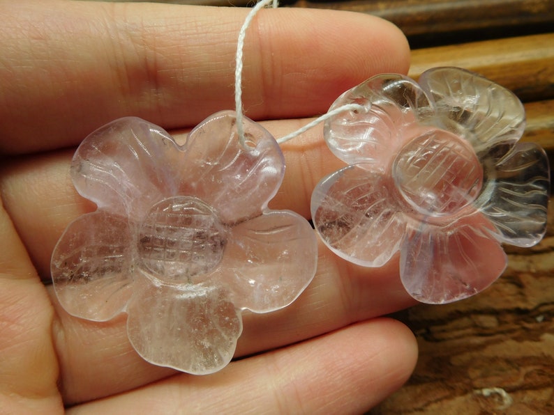 May include: Two clear, translucent flower-shaped pendants made of amethyst. The flowers have five petals and a center with a grid pattern. The pendants are strung on a white cord.