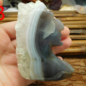 Puede incluir: Una piedra de &aacute;gata tallada en forma de perfil de un nativo americano. La piedra es de color gris claro con bandas blancas y azul claro.