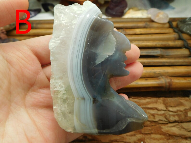 Puede incluir: Una piedra de &aacute;gata tallada en forma de perfil de un nativo americano. La piedra es de color gris claro con bandas blancas y azul claro.