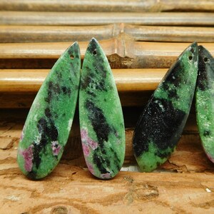 May include: Four elongated, polished gemstone pendants. The pendants are a mix of green, black, and red hues. Each has a hole drilled at the top for stringing. The background is a natural wood surface.