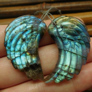 May include: Two iridescent blue labradorite gemstone wings carved in a detailed feather pattern. The wings are approximately 2 inches long and 1 inch wide.
