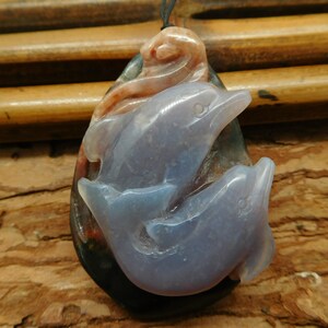 May include: A pendant carved from agate featuring two dolphins, one leaping over the other. The dolphins are carved in a light blue color, while the agate base is a mix of black, brown, and red.