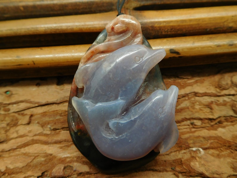May include: A pendant carved from agate featuring two dolphins, one leaping over the other. The dolphins are carved in a light blue color, while the agate base is a mix of black, brown, and red.