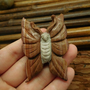 May include: A hand holding a carved butterfly made of brown and white banded stone. The butterfly has detailed wings and a white face.