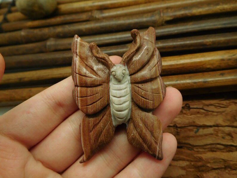 May include: A hand holding a carved butterfly made of brown and white banded stone. The butterfly has detailed wings and a white face.