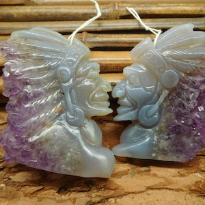 May include: Two carved amethyst and agate pendants in the shape of Native American chiefs' heads. The pendants are facing each other and have detailed carvings of feathers and faces.