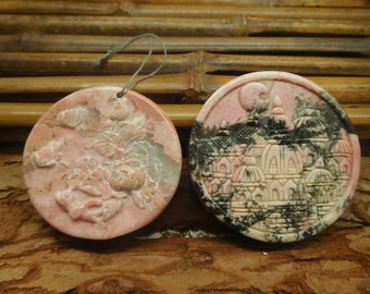 May include: Two round pink and black stone pendants with carved designs. One pendant features a floral design and the other features a cityscape.