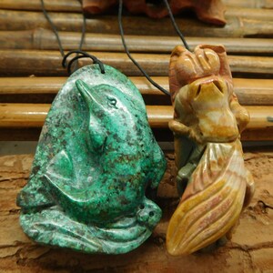 Puede incluir: Dos colgantes de piedra tallada en cordones negros. El colgante de la izquierda es verde y presenta un delfín. El colgante de la derecha es una piedra multicolor tallada en una figura estilizada.