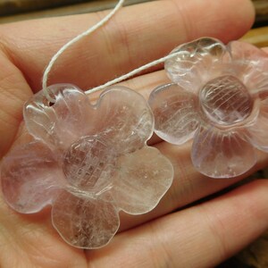 May include: Two clear, pale lavender flower-shaped pendants with a carved design in the center. The pendants are strung on a white cord.