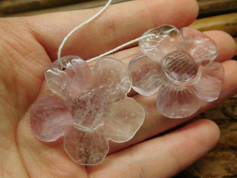 May include: Two clear, pale lavender flower-shaped pendants with a carved design in the center. The pendants are strung on a white cord.