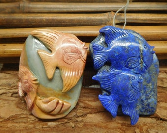 May include: Two carved stone pendants featuring fish designs. One pendant is a mix of peach, tan, and gray, while the other is a deep blue. The fish are intricately carved with detailed fins and scales, showcasing a natural theme.