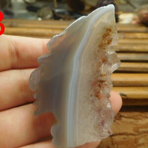 Puede incluir: Un cristal de &aacute;gata gris y morado con un borde natural y &aacute;spero. El cristal tiene una banda blanca que lo atraviesa y un grupo de cristales m&aacute;s peque&ntilde;os en un lado.