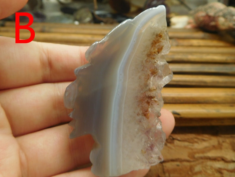 Puede incluir: Un cristal de &aacute;gata gris y morado con un borde natural y &aacute;spero. El cristal tiene una banda blanca que lo atraviesa y un grupo de cristales m&aacute;s peque&ntilde;os en un lado.