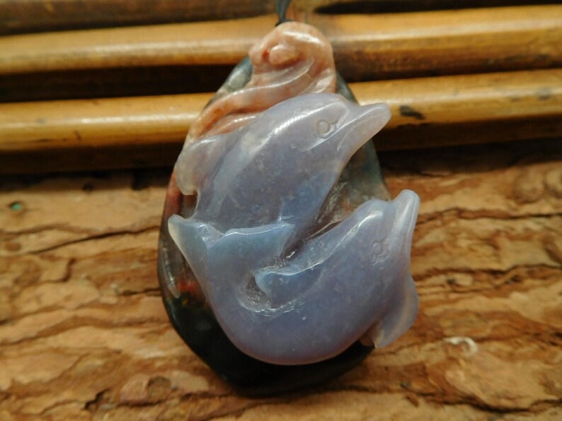 May include: A pendant featuring two carved dolphins in a light blue agate, set in a multi-colored agate stone. The pendant is teardrop shaped and has a hole at the top for a cord or chain.