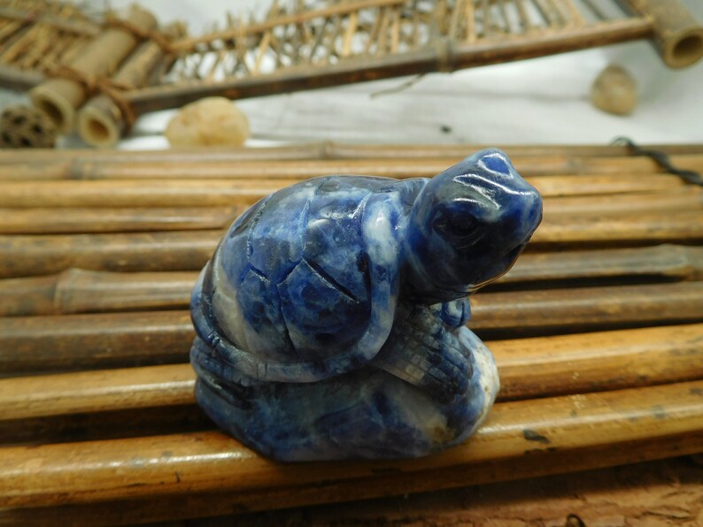 May include: A small blue lapis lazuli turtle sculpture. The turtle is carved in a sitting position with its head turned to the side. The sculpture is detailed with intricate carvings and has a natural, polished finish.