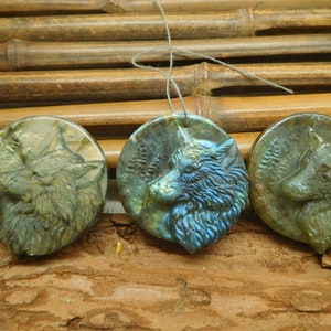 Puede incluir: Tres colgantes redondos tallados en piedra labradorita, cada uno con la cabeza de un lobo de perfil. El pelaje del lobo está detallado con un acabado texturizado, y la piedra tiene un brillo iridiscente azul verdoso.