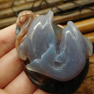 May include: A pendant carved from agate featuring two dolphins intertwined. The dolphins are a light purple color with a translucent quality. The pendant is set against a background of brown and pink agate.