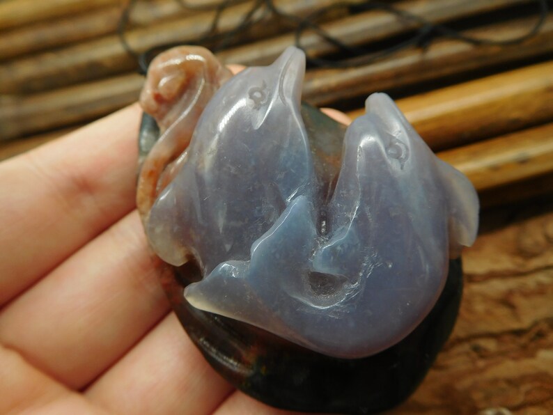 May include: A pendant carved from agate featuring two dolphins intertwined. The dolphins are a light purple color with a translucent quality. The pendant is set against a background of brown and pink agate.