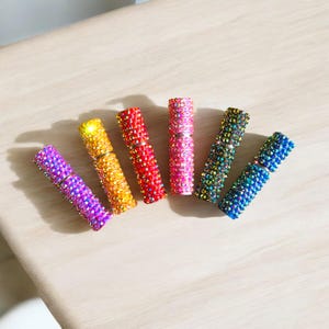 Könnte beinhalten: Sechs farbenfrohe Parfümzerstäuber, besetzt mit glitzernden Strasssteinen. Die Zerstäuber sind diagonal angeordnet und zeigen eine Vielzahl von Farben: Lila, Gelb, Rot, Rosa, Grün und Blau. Jeder Zerstäuber ist mit kleinen, funkelnden Strasssteinen bedeckt.