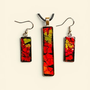 May include: A jewelry set consisting of a rectangular pendant on a black cord and two matching rectangular drop earrings. Each piece features a vibrant mosaic pattern of red, orange, and lime green fragments set within a dark metal frame.