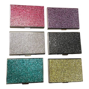 Op de afbeelding: Zes rechthoekige etuis, elk bedekt met sprankelende strass-steentjes. De etuis zijn in verschillende kleuren: felroze, paars, zilver, zwart, blauwgroen en goud. De etuis hebben een zilverkleurig metalen frame en een kliksluiting.