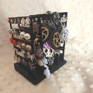 May include: A black, multi-tiered earring display stand filled with various earrings. The earrings include studs, hoops, and dangling styles in silver, gold, and pearl. Some earrings have unique designs, such as gears, skulls, and floral accents.