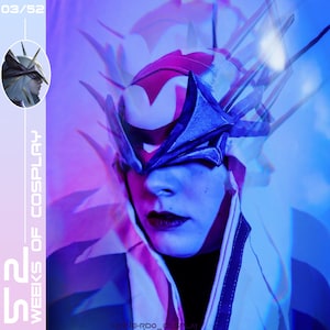 Puede incluir: Una persona con un disfraz azul y rosa con un tocado grande y detallado. El tocado está hecho de metal y tiene un diseño afilado y angular. El rostro de la persona está oculto por el tocado, pero sus labios son visibles y pintados de rojo oscuro. El fondo es un desenfoque de luz azul y rosa.