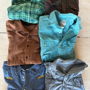 May include: Six different button-down shirts in various colors and patterns. The shirts are made of different materials, including denim, flannel, and cotton.