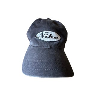 May include: A dark gray baseball cap featuring a white oval patch on the front panel. The word Nike is embroidered in black script lettering across the center of the white oval. The cap has a curved brim and visible stitching along the front.