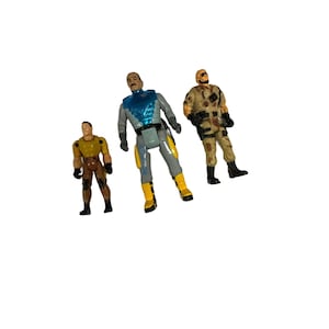 May include: Three plastic action figures, one in a blue and gray suit, one in a brown camouflage uniform, and one in a brown and yellow uniform.
