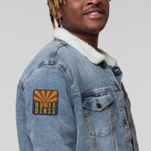 May include: A light blue denim jacket with a sherpa collar and a patch on the left sleeve. The patch is red, yellow, and blue with the text "NUNYA BZNSS".