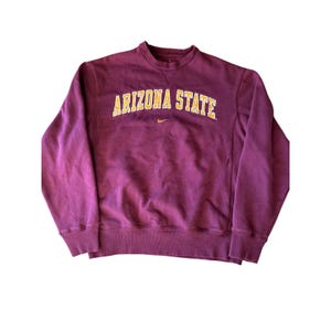 May include: A maroon Arizona State University sweatshirt with the words "ARIZONA STATE" in gold lettering across the chest. A small gold Nike swoosh is visible below the text. The sweatshirt has a crew neck and ribbed cuffs.