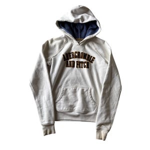 May include: Cream-colored Abercrombie and Fitch hoodie with a navy blue lined hood. The hoodie features a kangaroo pocket and the brand name in a brown and gold arched design across the chest. The hoodie has a drawstring and ribbed cuffs.