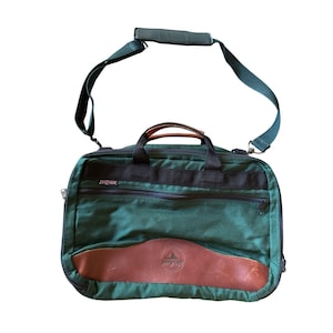 May include: Green JanSport briefcase with a brown leather accent and a black top handle. The bag has a detachable green shoulder strap and a zippered front pocket. The JanSport logo is visible on the front.
