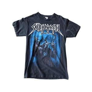 May include: Black t-shirt featuring the band name "SKELETONWITCH" in white, stylized lettering above a graphic of a hooded figure with a scythe. The graphic is set against a blue background.