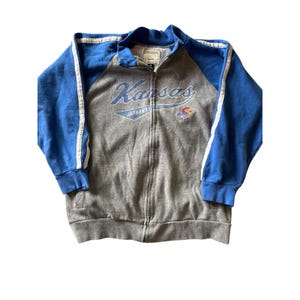 May include: A gray zip-up jacket with blue sleeves and white stripes. The jacket features the word "Kansas" in blue script across the chest, with "JAYHAWKS" below. A Jayhawk logo is on the right side.