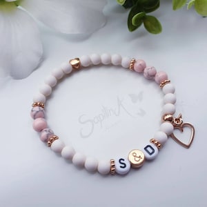 Könnte beinhalten: Armband mit weißen und rosa Perlen und goldenen Akzenten. Das Armband hat weiße und rosa Perlen, goldfarbene Abstandshalter und einen goldenen Herzanhänger. Die Buchstaben "S & D" sind auf dem Armband zu sehen.