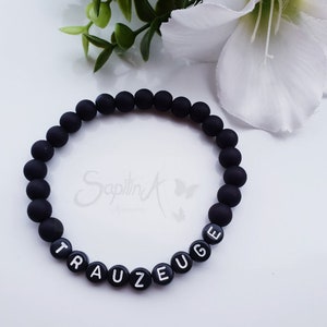 May include: Black beaded bracelet with the word "TRAUEUGE" spelled out in white letters.