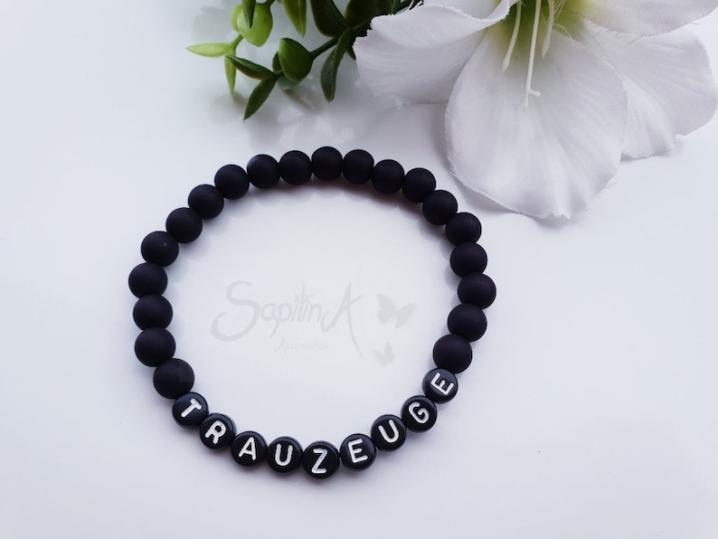 May include: Black beaded bracelet with the word "TRAUEUGE" spelled out in white letters.