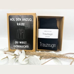 May include: A brown box with a white label that says "Hol den Anzug raus!" and "Du wirst gebraucht!" with a black and white illustration of a suit. The box contains a pair of black socks with a white label that says "Wilst du mein Trauzeuge werden?" and "Trauzeuge".
