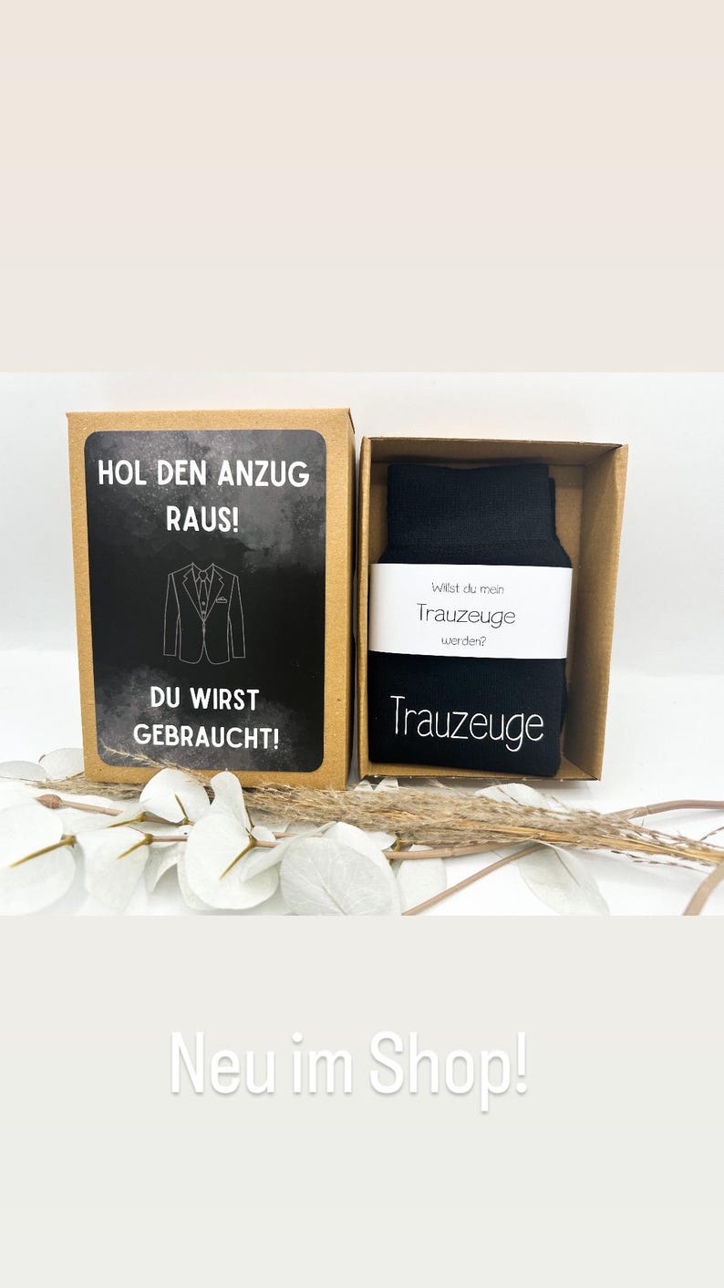 May include: A brown box with a white label that says "Hol den Anzug raus!" and "Du wirst gebraucht!" with a black and white illustration of a suit. The box contains a pair of black socks with a white label that says "Wilst du mein Trauzeuge werden?" and "Trauzeuge".