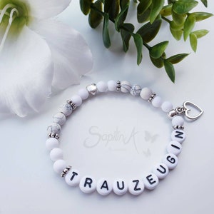 Könnte beinhalten: Weißes Perlenarmband mit einem silbernen Herzanhänger und dem Wort "TRAUZEUGIN" in schwarzen Buchstaben auf den Perlen.