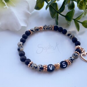 Könnte beinhalten: Ein Perlenarmband mit schwarzen, gesprenkelten und goldfarbenen Perlen. Das Armband hat goldene Herzanhänger und Buchstabenperlen mit der Aufschrift "VS". Das Armband ist mit einem elastischen Band versehen.