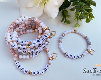 May include: A set of white and pink beaded bracelets with gold accents. The bracelets spell out "Team Bride" and "Bride".